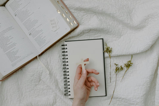 Hand holding wooden cross over notebook beside open Bible and dried flowers on white quilt faith based gifts for women